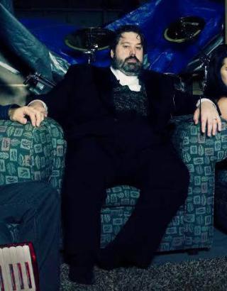 Joel Corda in a suit on a green chair in front of a blue wall