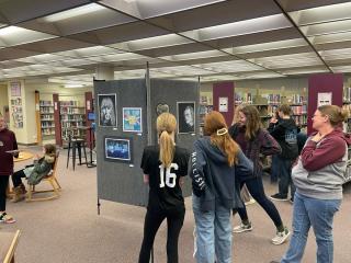 Youth Art on display in library, three teenage girls looking at paintings on gray pallets. 
